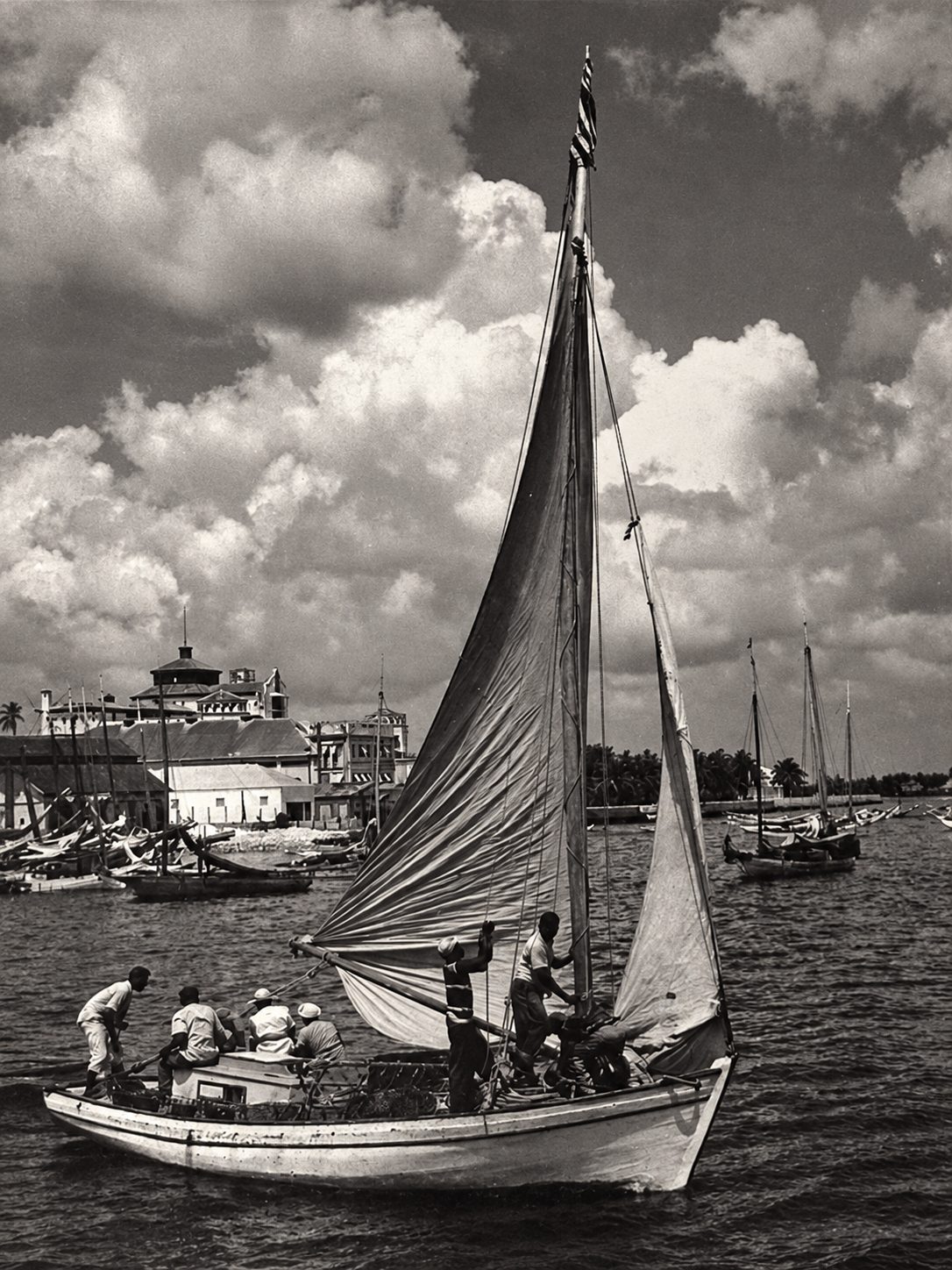 An archival black-and-white photograph of several men crewing a sailboat, with a distinctive harbourside building visible in the background