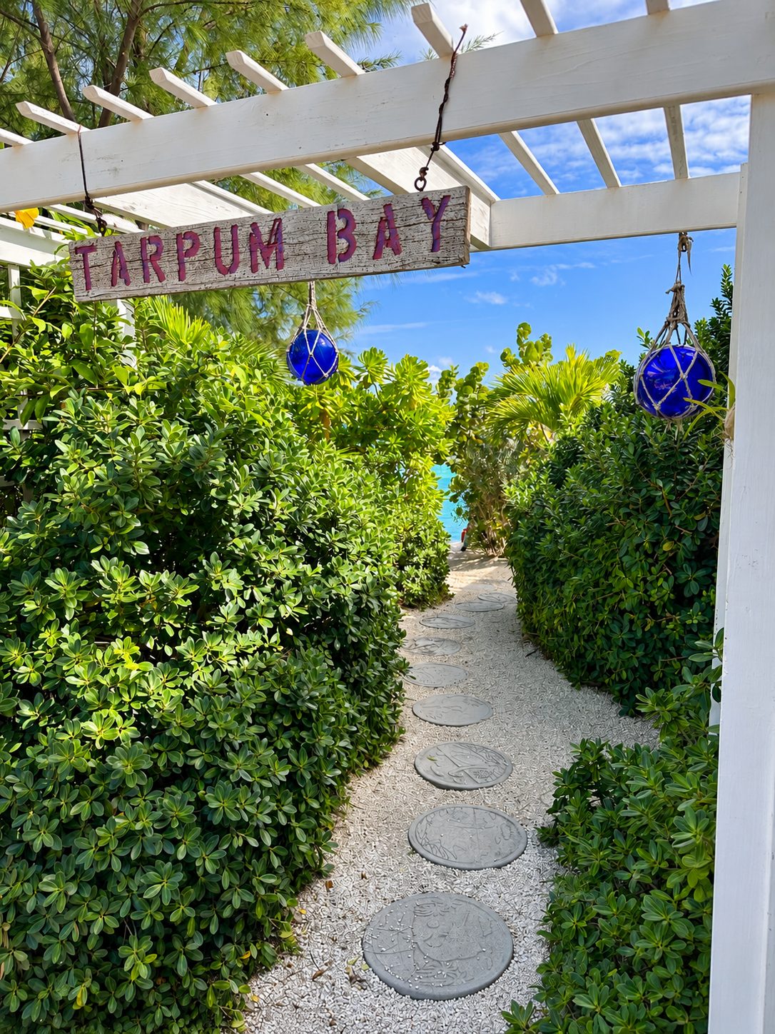 A wooden garden sign reading Tarpum Bay at the entrance to a pergola, with blue glass fishing floats hanging nearby and a stepping-stone path leading toward a view of the sea